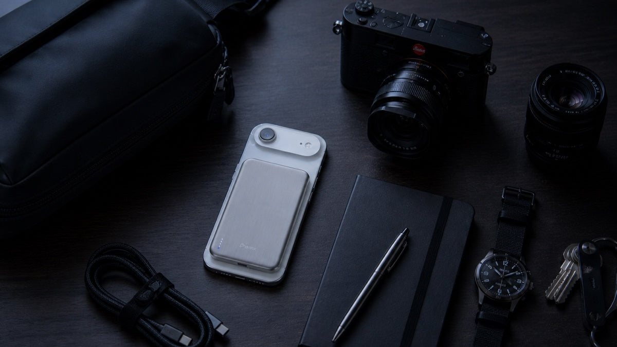 BMX SolidSafe Air 5K silver power bank magnetically snapped to a phone on a dark wooden desk, surrounded by everyday carry essentials: Leica camera, extra lens, automatic watch, keys, notebook, pen, USB-C cable, and dop kit